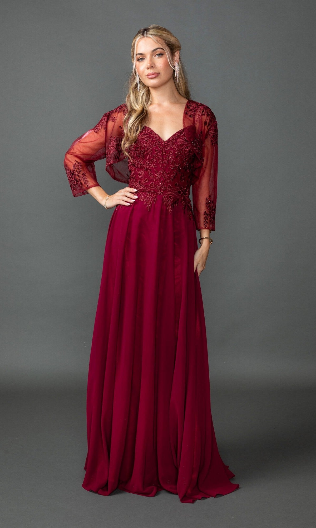 A woman stands before a gray backdrop wearing the Dancing Queen 4542, a long burgundy prom dress with lace embroidery on sheer sleeves and a fitted bodice.